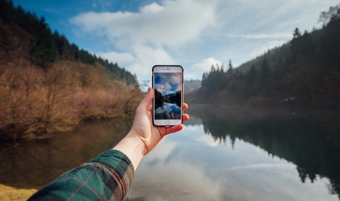 Man in plaid shirt holds phone out to take a photo of a lake with trees in autumn