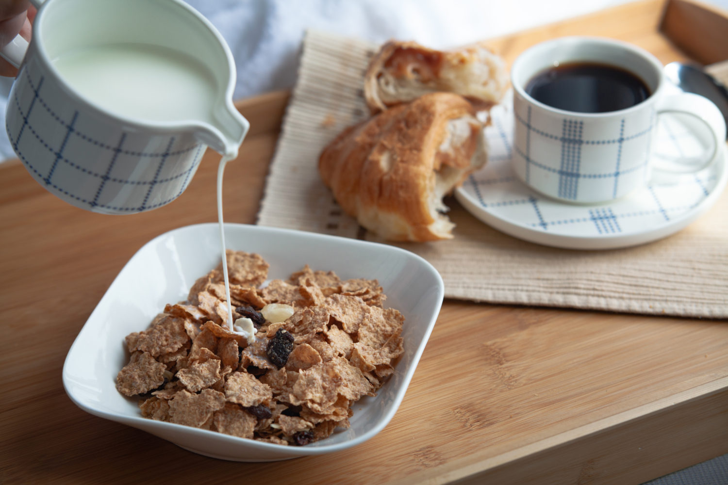 A small container of milk pouring into cereal next to a croissant and coffee