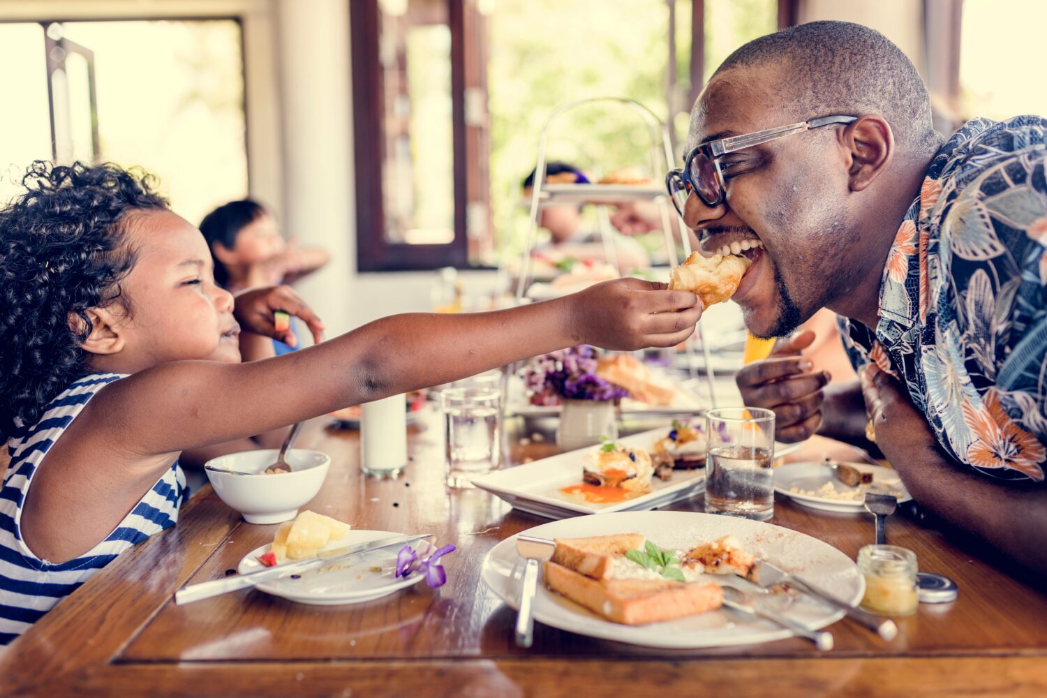 young daughter reaching across the table to feed her father breakfast