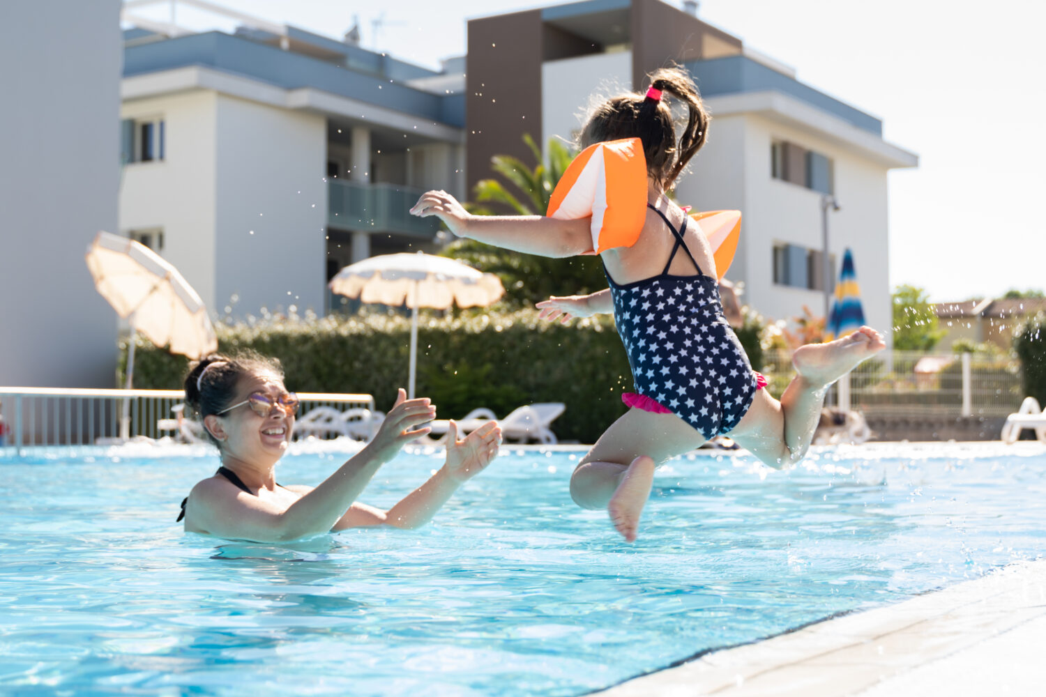 child jumping into a pool wearing floaties, and their mom waiting with open arms