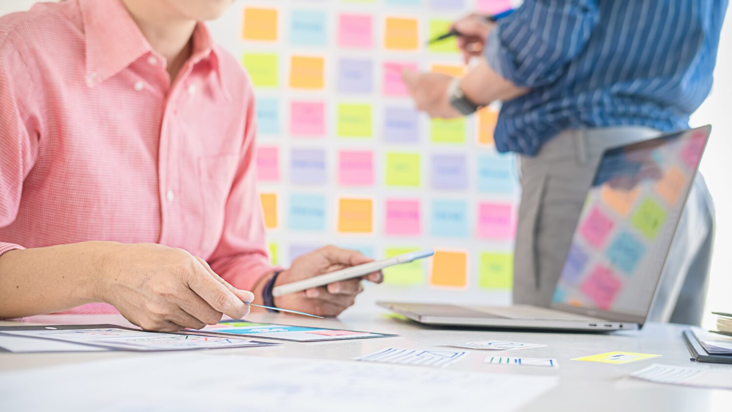 foreground business person looking at a laptop and background business person adding sticky notes to the wall