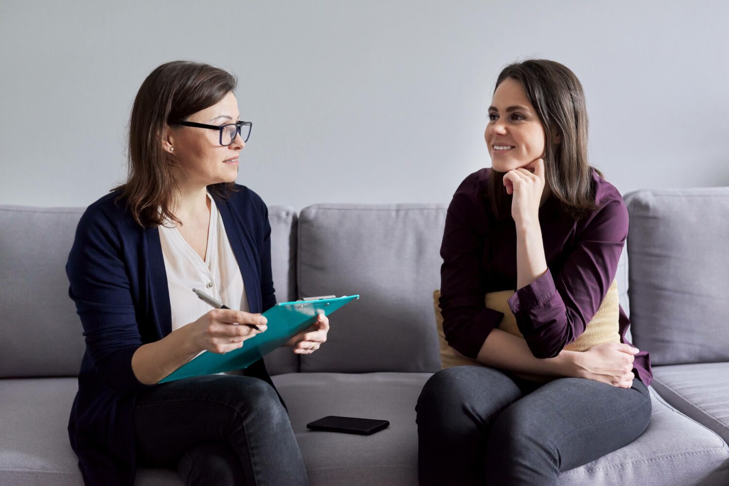 two women sitting on a couch and talking, woman on the left holding a clipboard