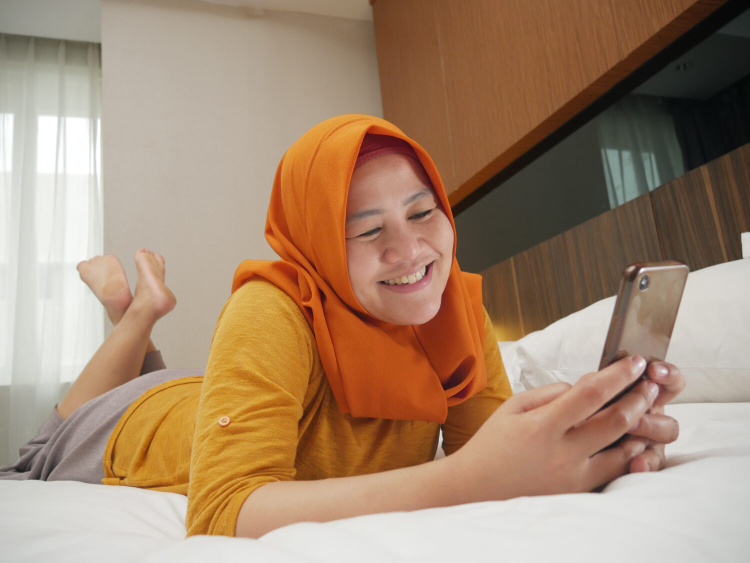muslim asian woman in orange hijab laying on hotel bed texting happily