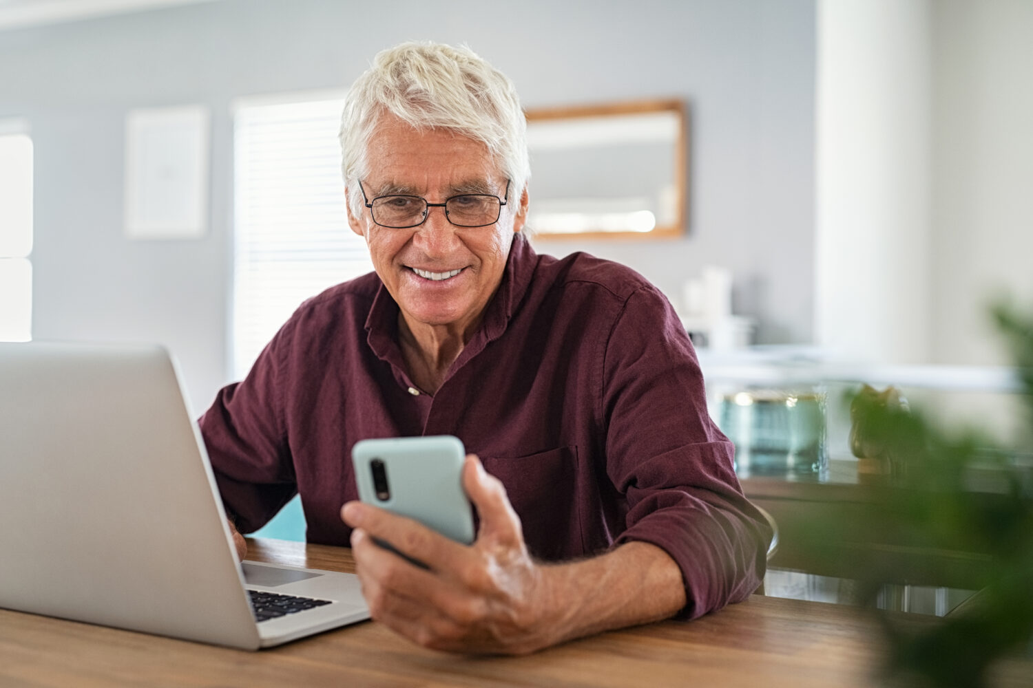older male working at a laptop and smiling down at his phone