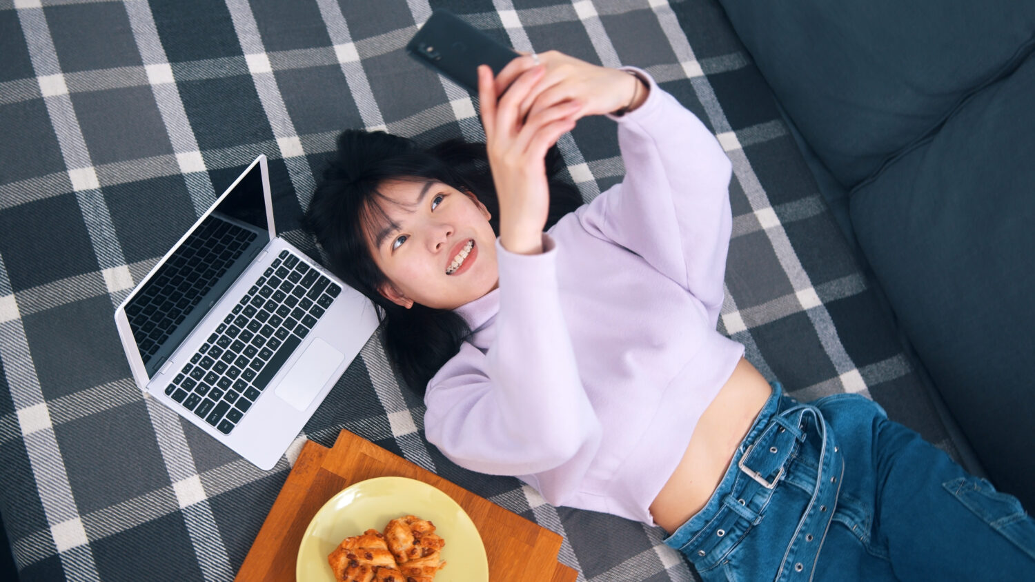 young asian woman laying on a bed beside her computer and taking a selfie