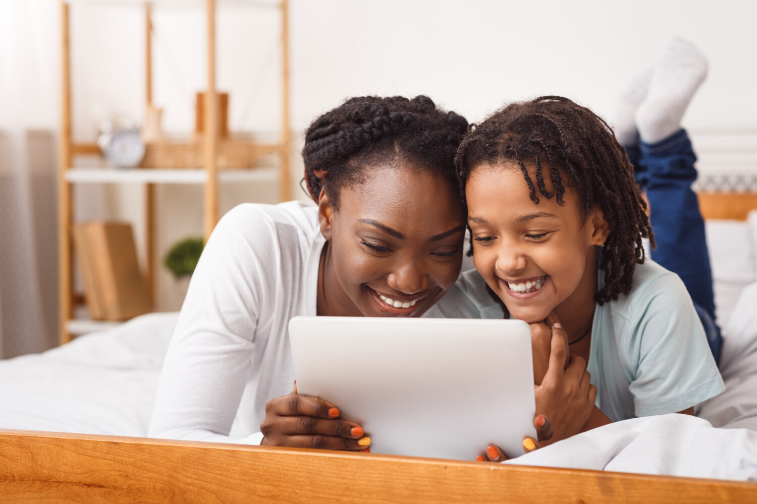 black mother and daughter laying in bed and smiling at a white tablet