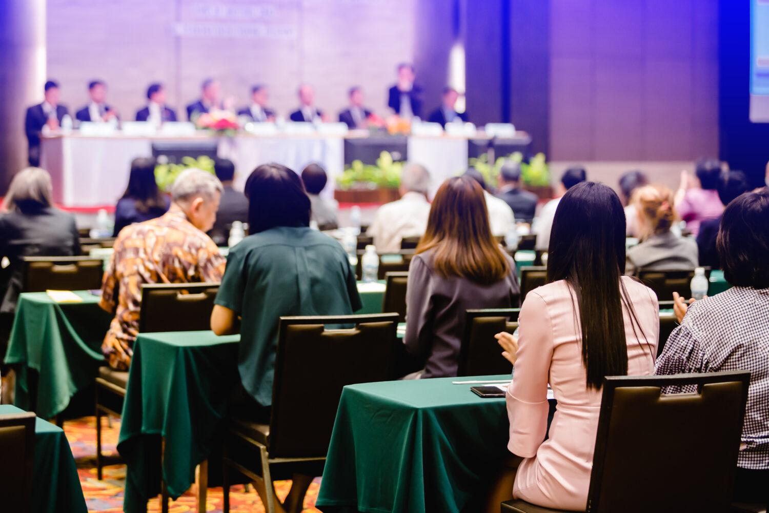 conference room tables with green tablecloth and people sitting at them listening to a panel presentation